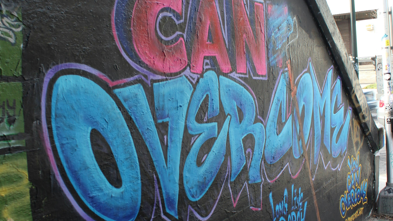 Colorful graffiti on a wall reading "CAN OVERCOME" in blue and red, with additional tags and artwork on a dark background.