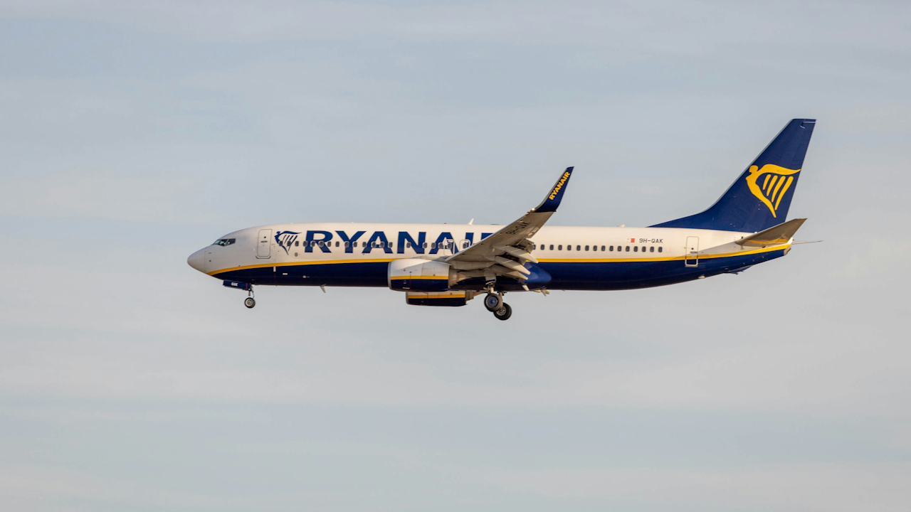 A white and blue Ryanair Boeing 737 airplane with its landing gear down is flying low against a light blue, cloudy sky.