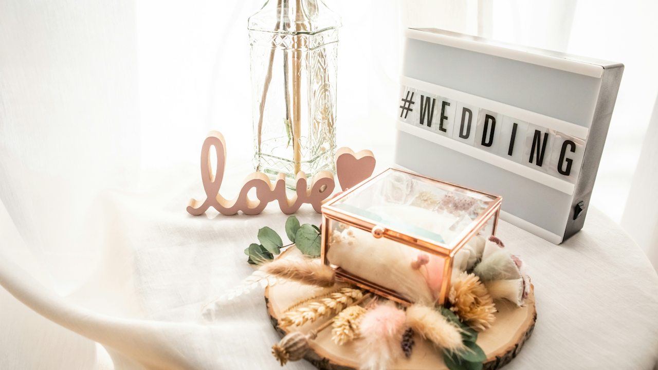 Wedding decor with "#WEDDING" sign, "love" letters, glass box with flowers, and vase on a wooden base.
