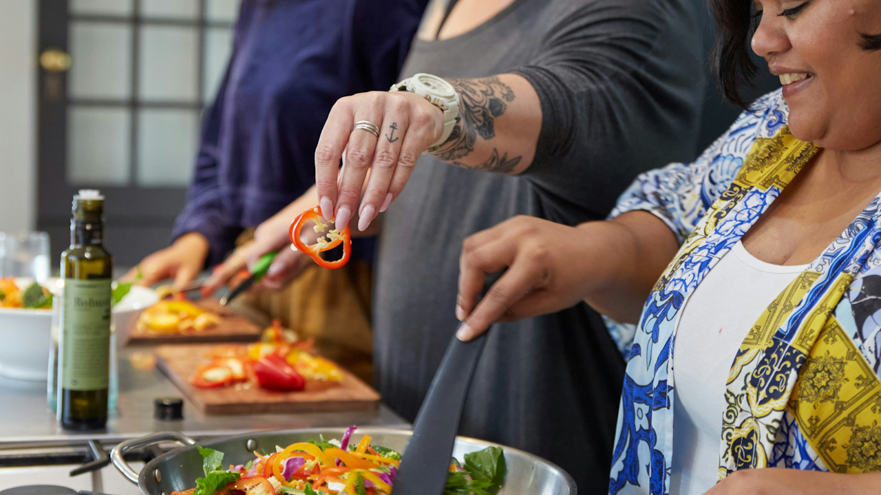 A group of people cooking together indoors, wearing various clothing styles and interacting while preparing food. Some are handling bottles and ingredients.