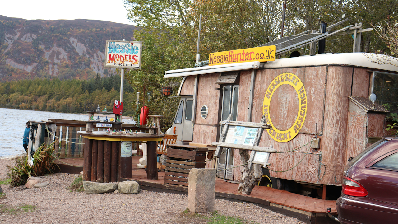 A small wooden trailer parked outdoors, featuring a table and a sign that reads "Nessie Models Since 1991". The scene includes trees, a clear sky, and the backdrop of mountains and a lake.