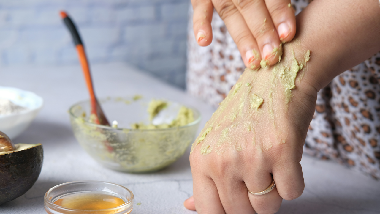A person's hand is shown applying a green, mashed avocado mixture to the back of another person's hand. In the background are ingredients like an avocado half and a small bowl of honey.
