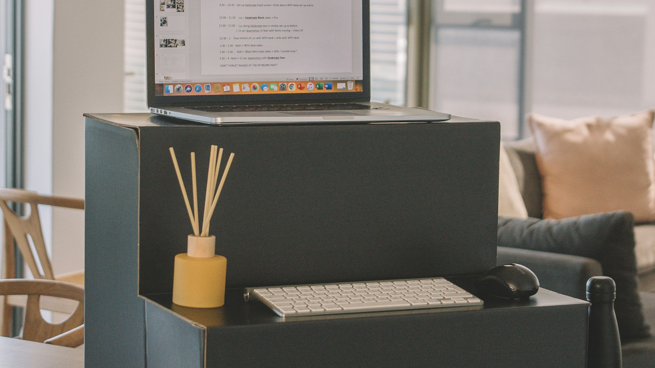 Laptop on a black stand with a white keyboard, mouse, and yellow diffuser, in a modern room with a chair and sofa.