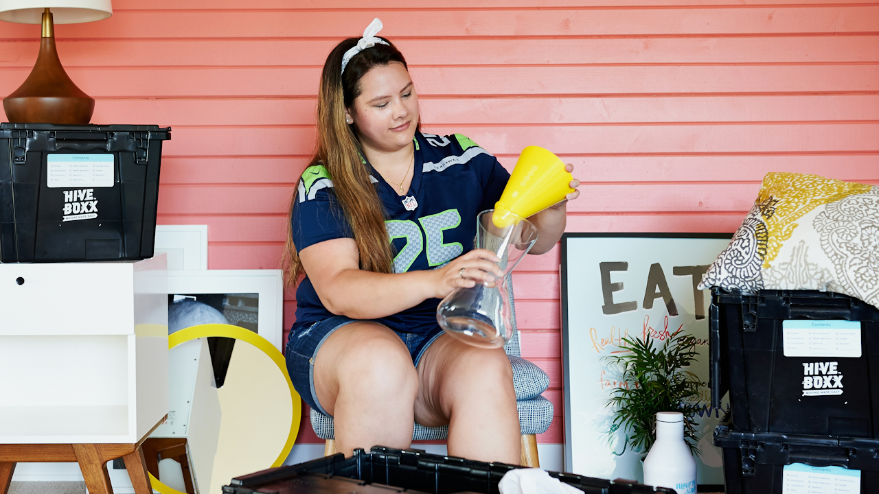 A woman with a smile is pouring a liquid into a beaker while sitting indoors, surrounded by furniture and a wall. She is wearing casual clothing.
