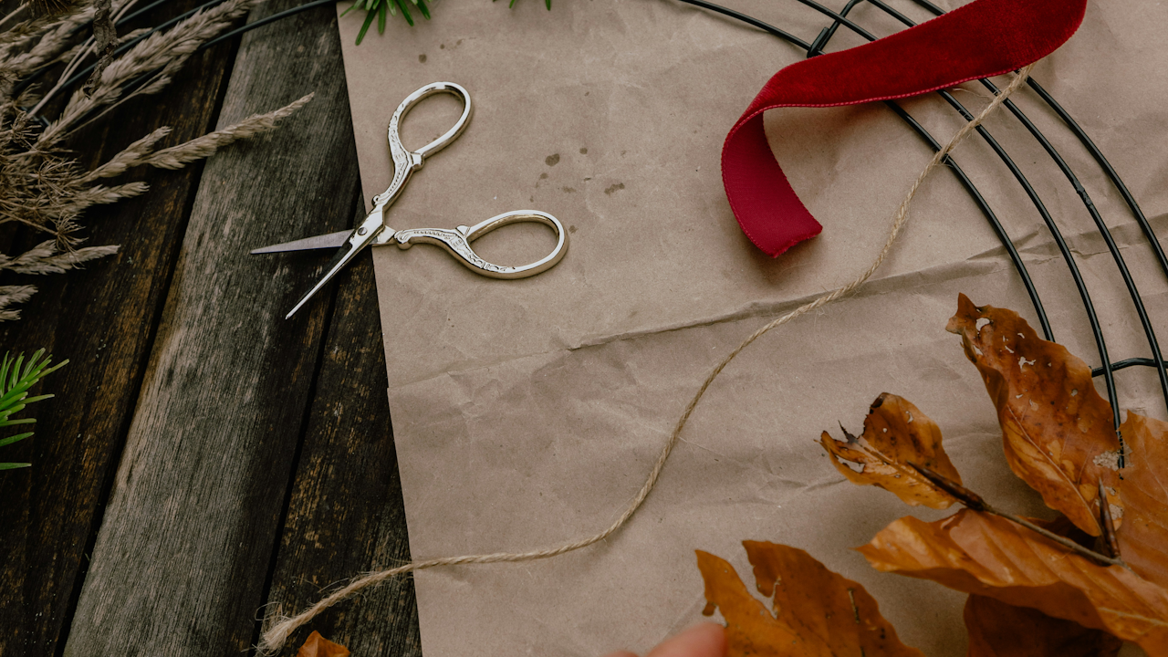 Crafting setup: ornate scissors, red ribbon, twine, wire wreath frame, dried leaves, and pine sprigs on rustic wood.