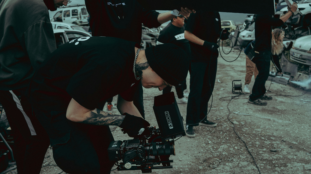 A man with tattoos on his arm is focused on working with a camera, dressed casually and standing on the ground.