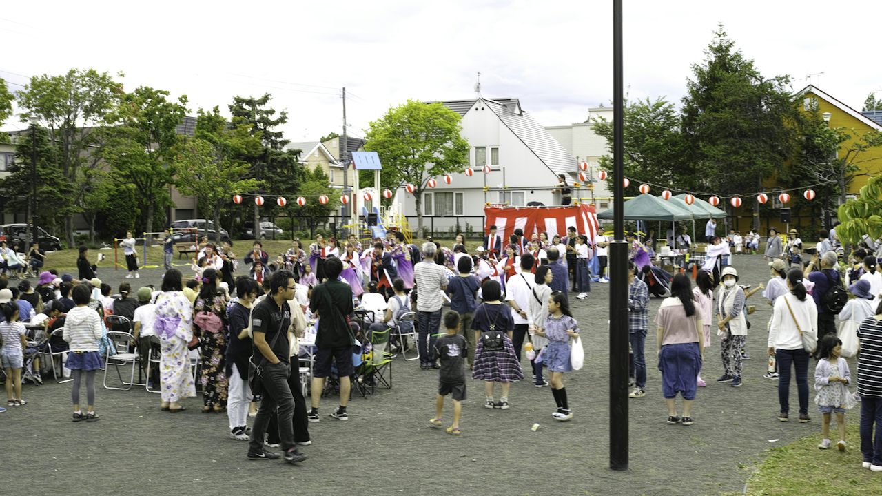 Crowded outdoor festival with people, traditional performers, red tents, lanterns, and houses in the background.