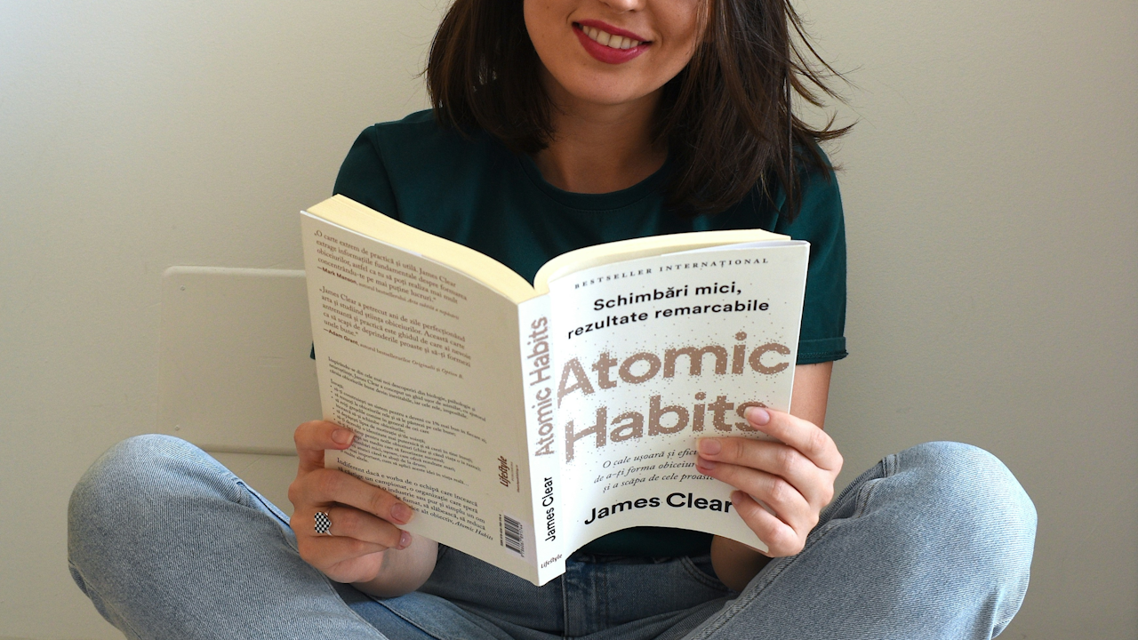 A smiling person sits cross-legged, holding an open copy of the book "Atomic Habits" by James Clear.