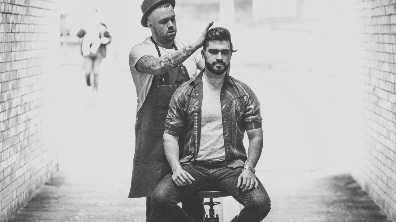 Barber cutting a man's hair in a black-and-white photo, set in a narrow brick alley with a blurred background.