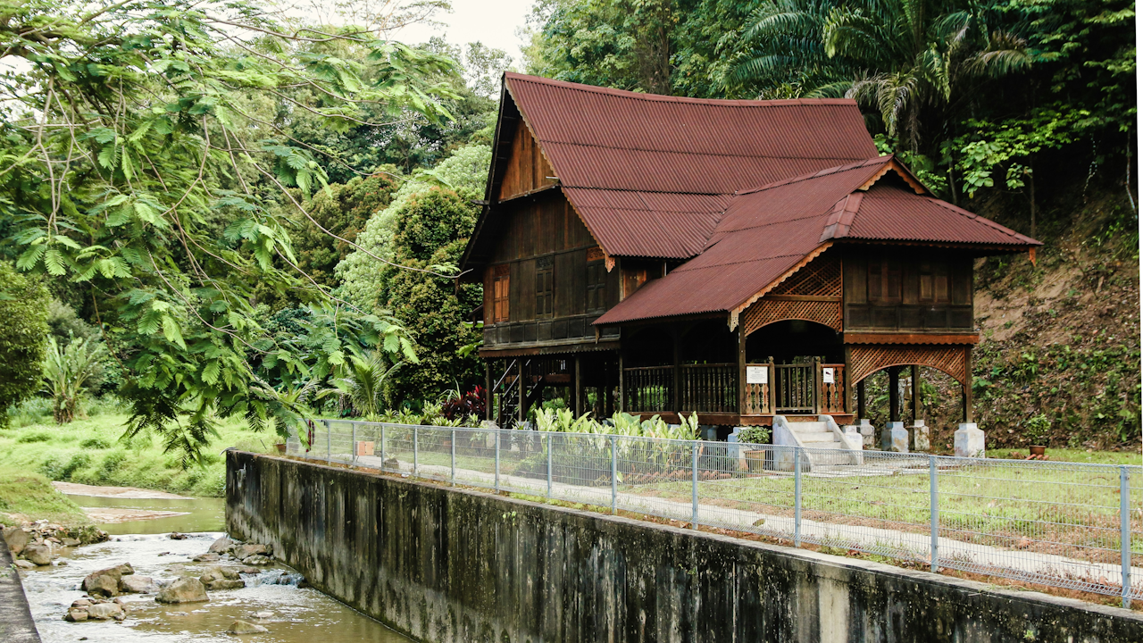 A house with a red roof situated beside a river, surrounded by trees and plants, featuring elements of Chinese and Japanese architecture.
