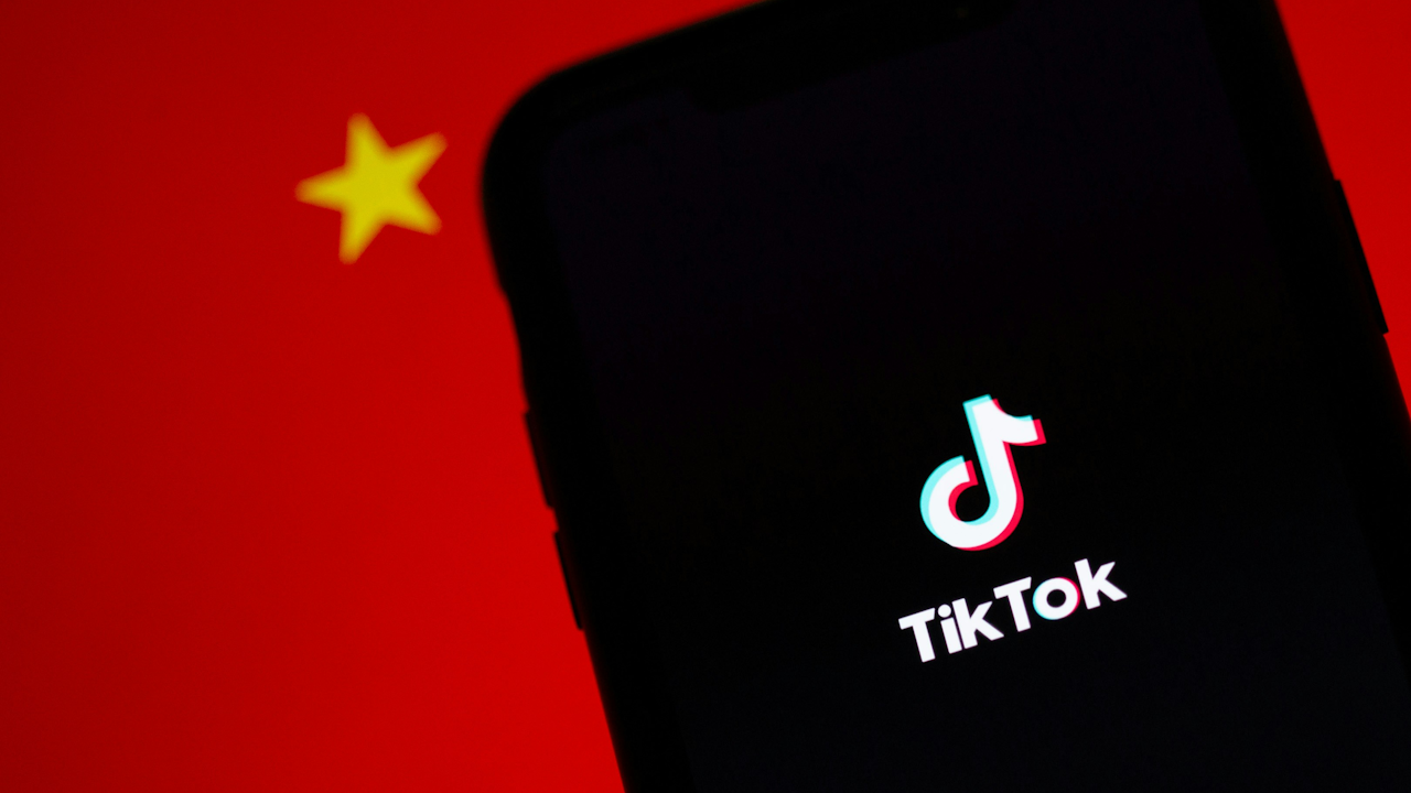 A smartphone screen displaying the TikTok logo and name. In the background, a part of the flag of the People's Republic of China is visible.