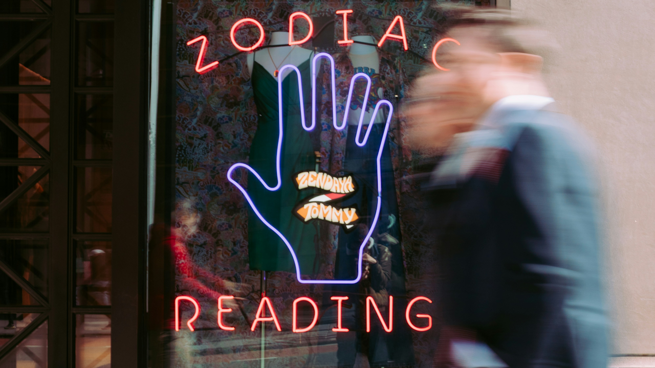 A blurry photo of a neon sign that says "ZODIAC READING" with a hand, seen through a storefront window with clothes on mannequins.