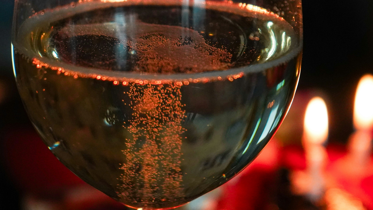 Close-up of sparkling white wine in glass, bubbles rising, blurred candles in warm background.