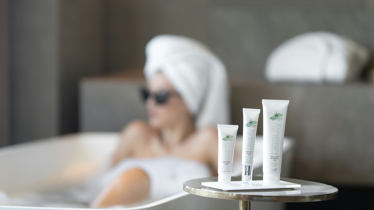 A person in a bathtub with a towel on their head, next to a table with three beauty cream tubes.