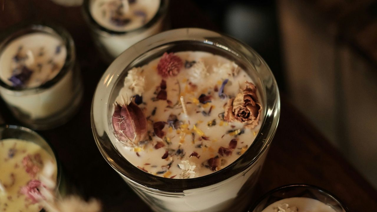 A glass candle with dried flowers embedded in white wax, surrounded by similar candles on a wooden surface.