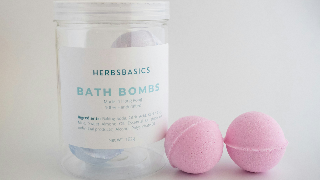 A jar of HerbsBasics bath bombs with two pink bath bombs next to it, made in Hong Kong with natural ingredients.