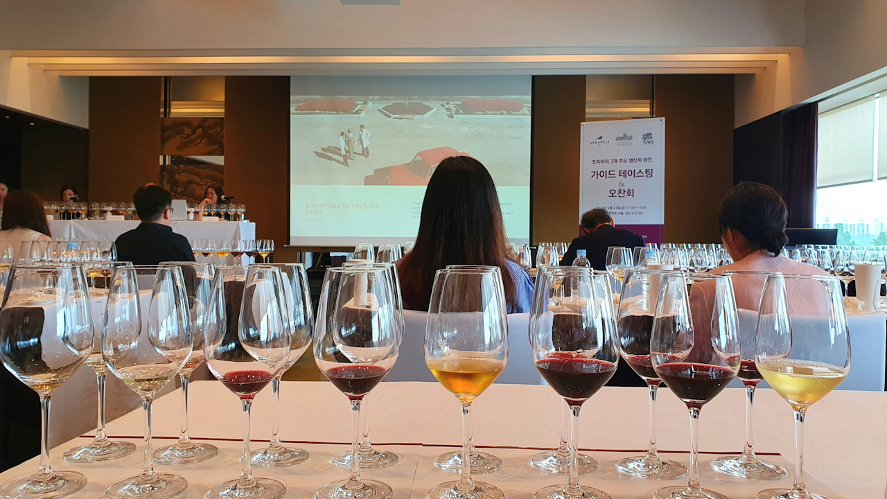 A first-person perspective image of a wine tasting event, with many glasses of wine lined up in the foreground, and people seated at tables in the background.