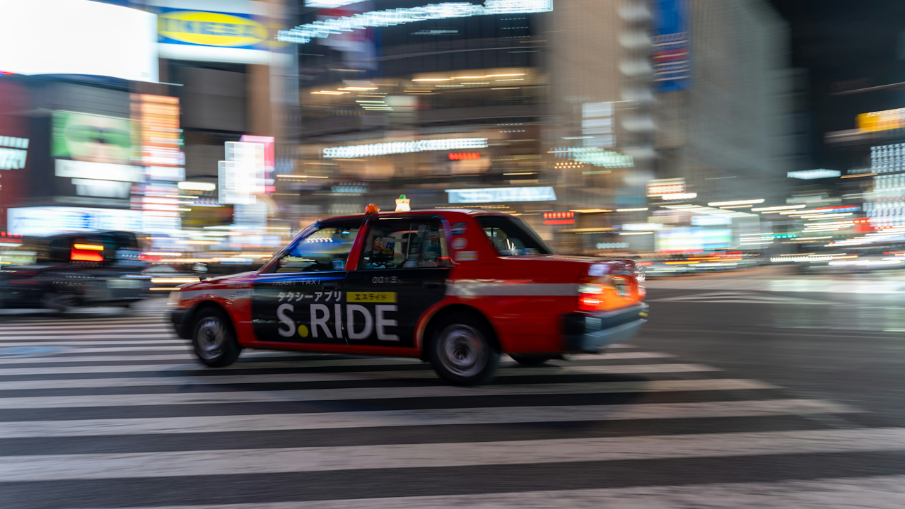 A red car driving on a street with buildings in the background.