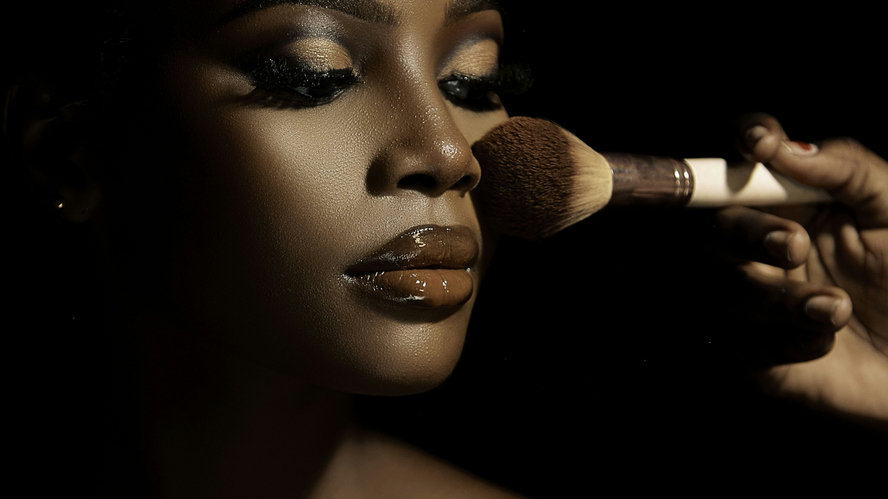 A close-up side profile of a person's face shows them getting makeup applied. A hand holds a large makeup brush, gently dusting powder onto their cheek. The person's eyes are closed, with long, defined eyelashes and shimmery gold eyeshadow. Their lips are glossy with a brown-toned lip gloss. The background is completely dark, highlighting the face and the makeup application.