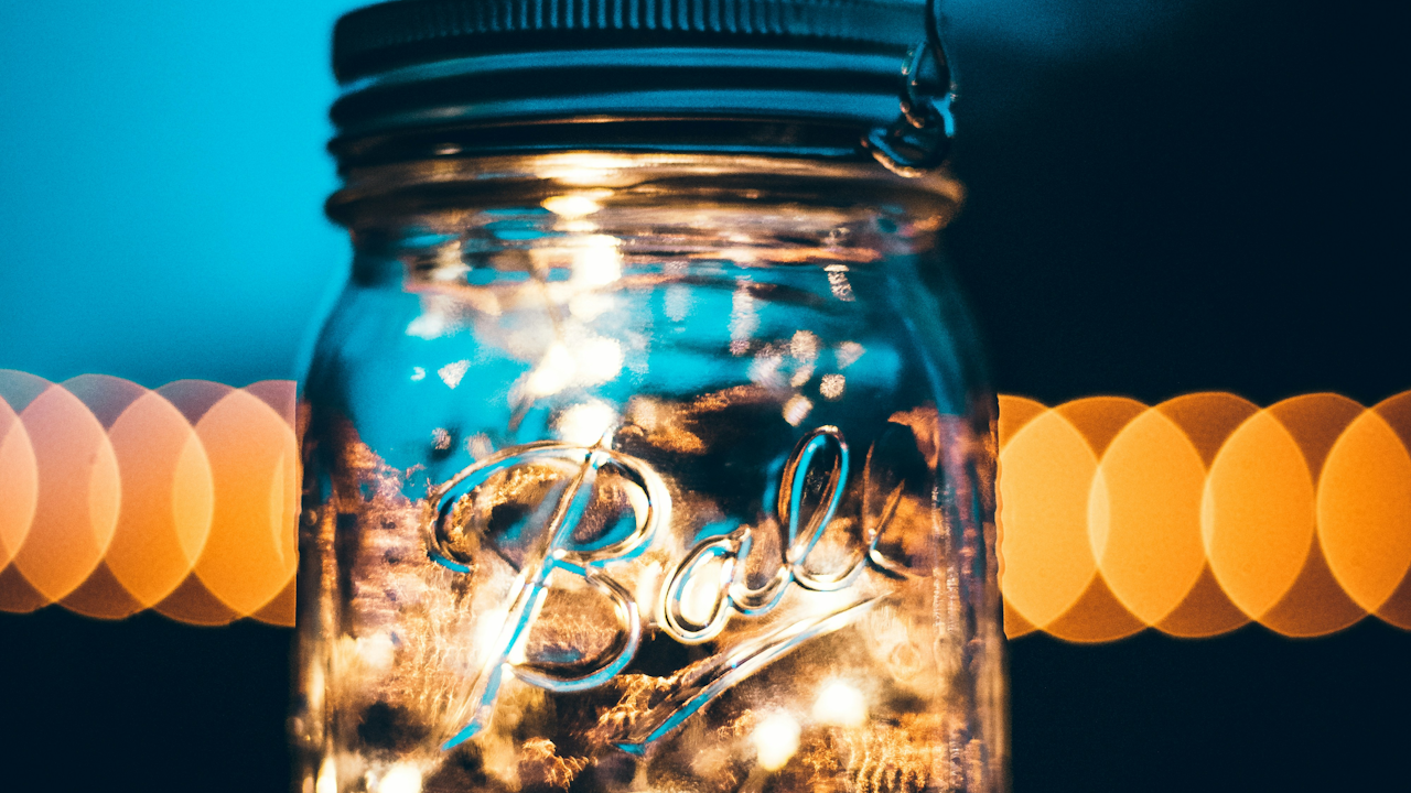 A glass jar filled with tiny, glowing lights, with a blurred background of warm, circular lights against a dark blue sky.