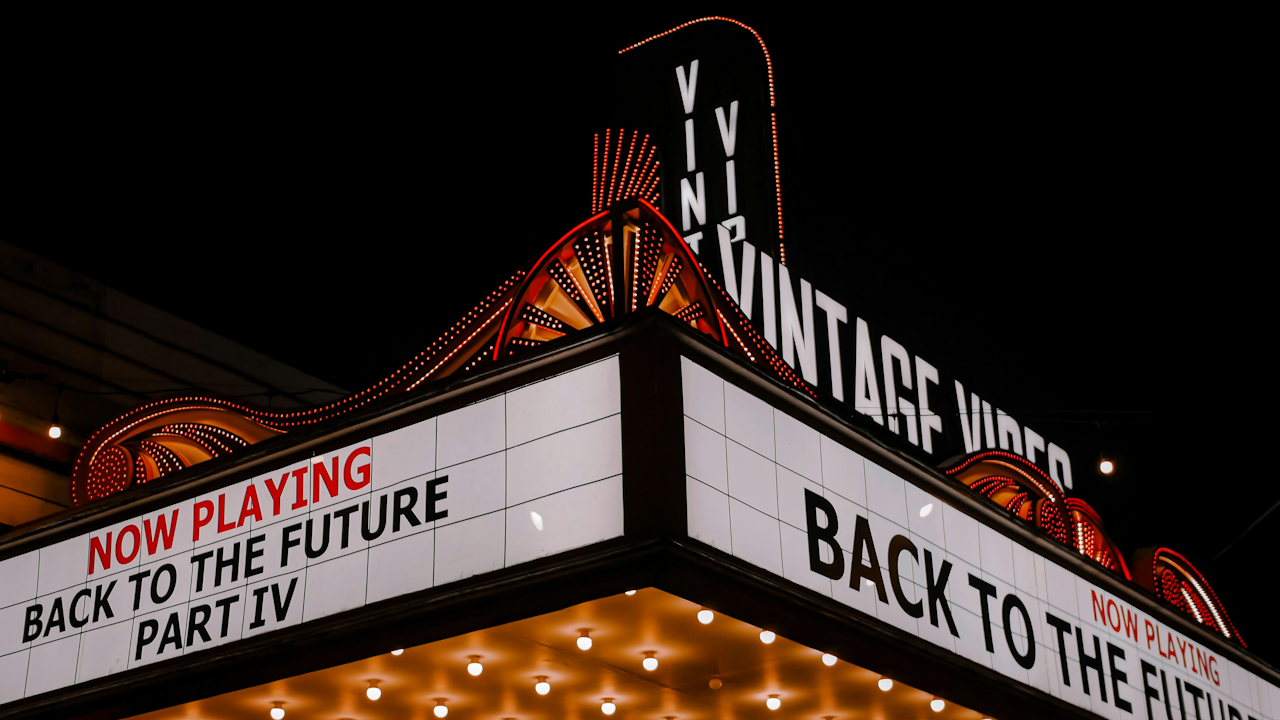 An electronic neon sign on a building displaying the text "NOW PLAYING BACK TO THE FUTURE" at night. The sign is illuminated and surrounded by the night sky, indicating it is part of a shop or business, possibly a theater or hotel.