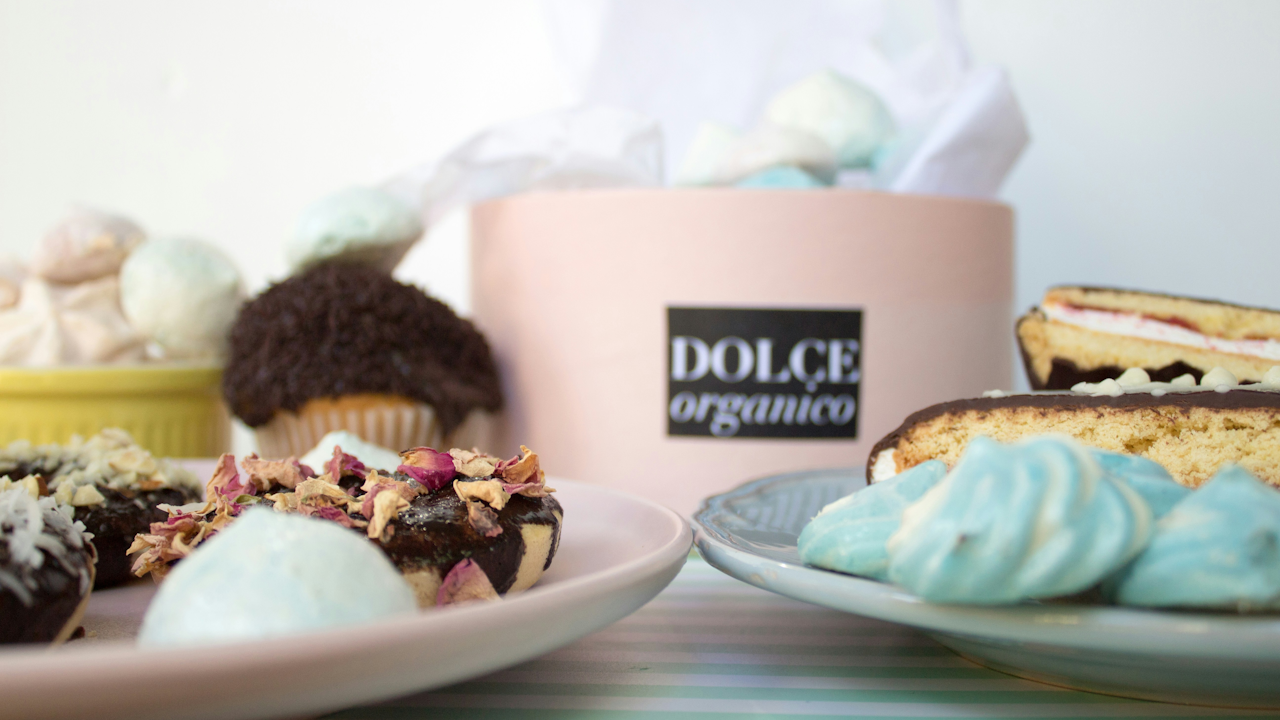 A close-up image of a delicious dessert featuring DOLCE organico. The focus is on a beautifully decorated baked good, possibly a cupcake or petit four, showcasing smooth buttercream icing, intricate cake decorating techniques, and vibrant food coloring. The dessert sits elegantly on a plate, surrounded by hints of other sweet treats characteristic of baking.