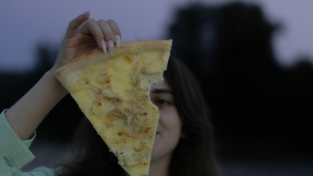 A woman standing outdoors holds a slice of pizza, ready to take a bite.