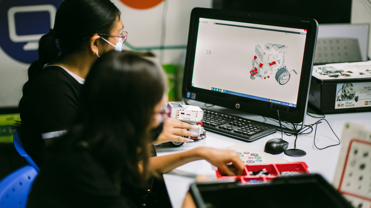 Students building robot with LEGO pieces at desk, computer showing robot design in classroom setting.