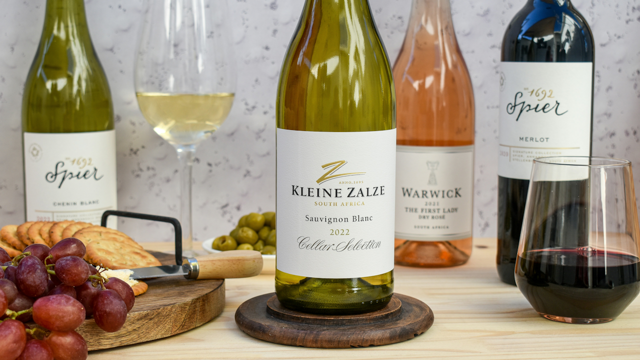 A variety of South African wines white, rosé, and red are displayed on a table with a wine glass, grapes, cheese, olives, and crackers.