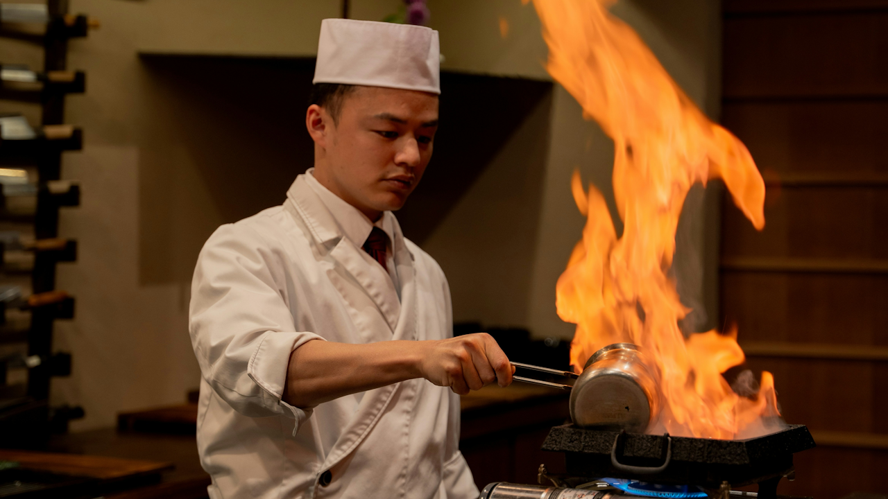 Chef in white uniform cooking with flames from a pan, surrounded by wine racks in a cozy kitchen.