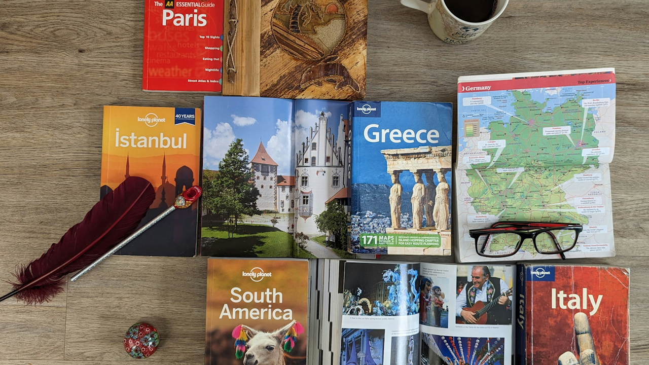 A wooden table is covered with various travel guides for destinations like Paris, Istanbul, Greece, South America, and Italy, along with a pair of glasses and a coffee mug.