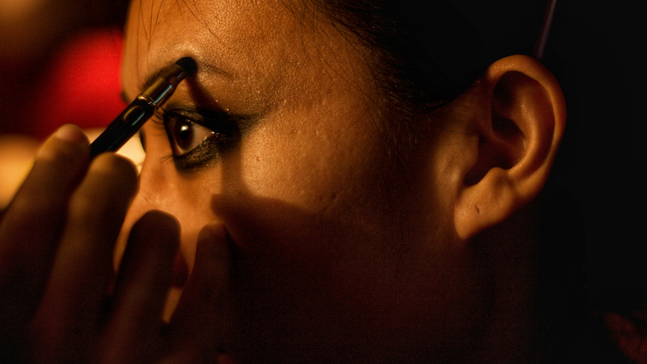 A close-up of a woman applying black eyeliner, showcasing her eyelashes and skin. The image focuses on her forehead and human face within an indoor setting.