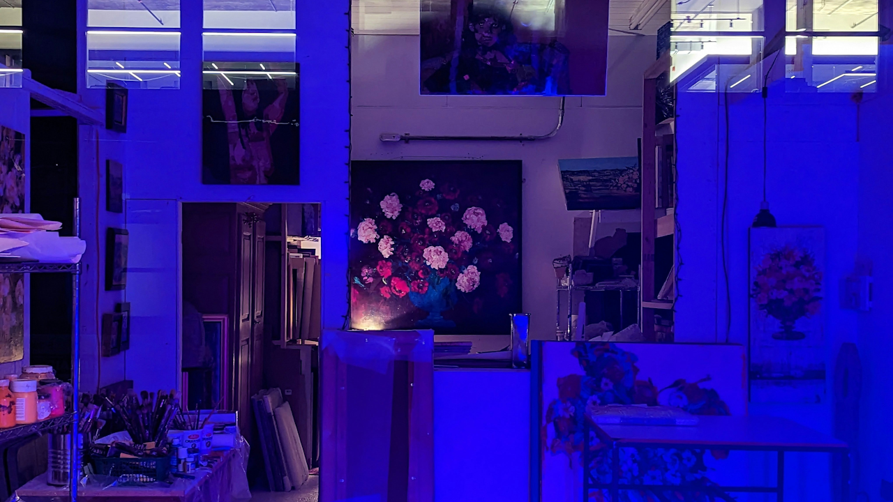 A studio is lit by a bright blue light, showcasing various paintings on the walls and easels, including a large floral piece.