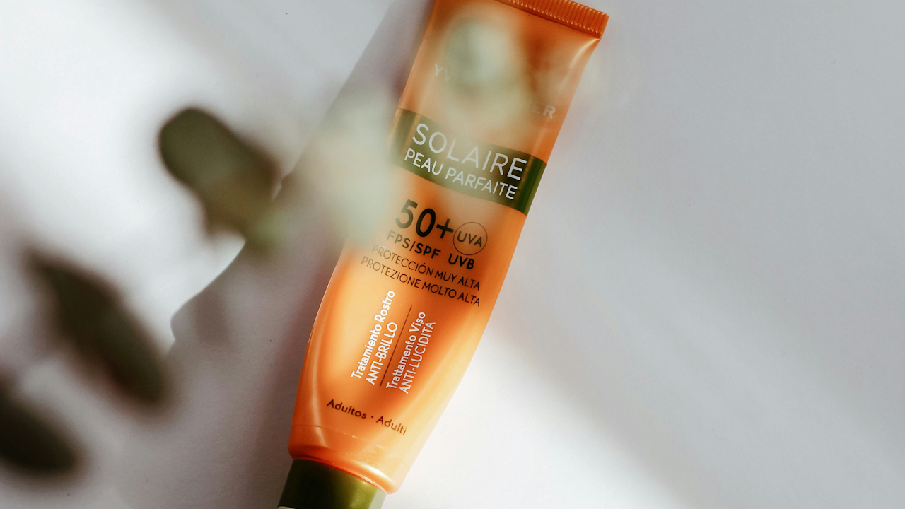 Orange sunscreen tube labeled "Solaire Peau Parfaite" with SPF 50+ UVA/UVB protection, on a white surface with blurred hand.