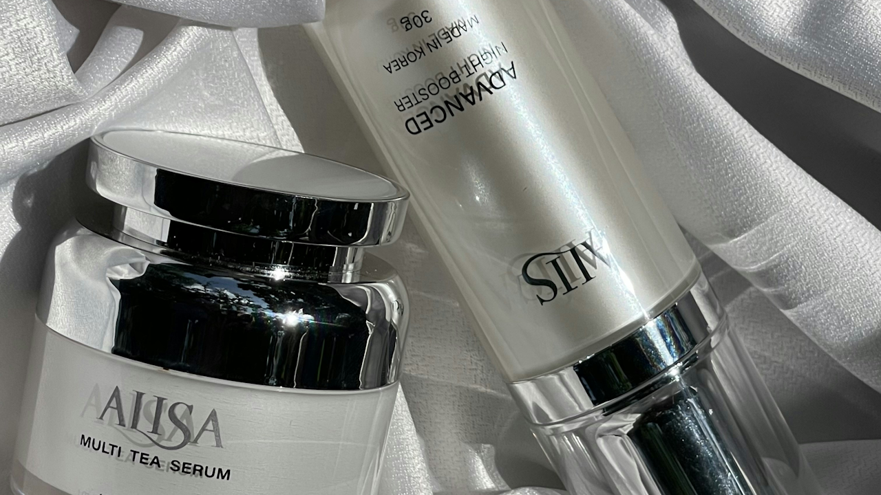 A high-angle close-up shot of two skincare bottles from the brand AALISA on a white satin cloth. The product names "MULTI TEA SERUM" and "ADVANCED NIGHT BOOSTER" are visible.