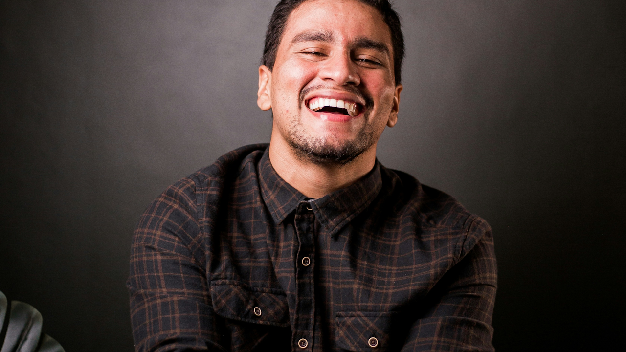 A man with a beard laughs with his mouth open, revealing his teeth. He is indoors against a wall, wearing a shirt. His expression shows joy and amusement.