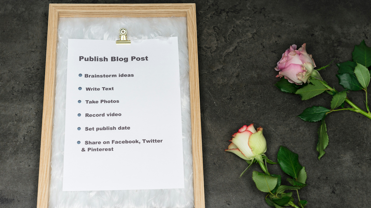 A top-down view of a framed checklist titled "Publish Blog Post" with six steps, next to two pink and white roses on a gray background.
