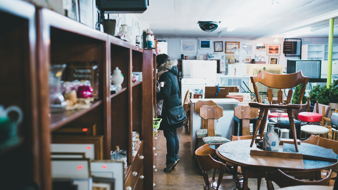 A person stands in a room with a shelf and several chairs, surrounded by furniture commonly found in dining areas, including a coffee table and countertop. The interior design reflects a casual atmosphere typical of a cafe or coffee shop.