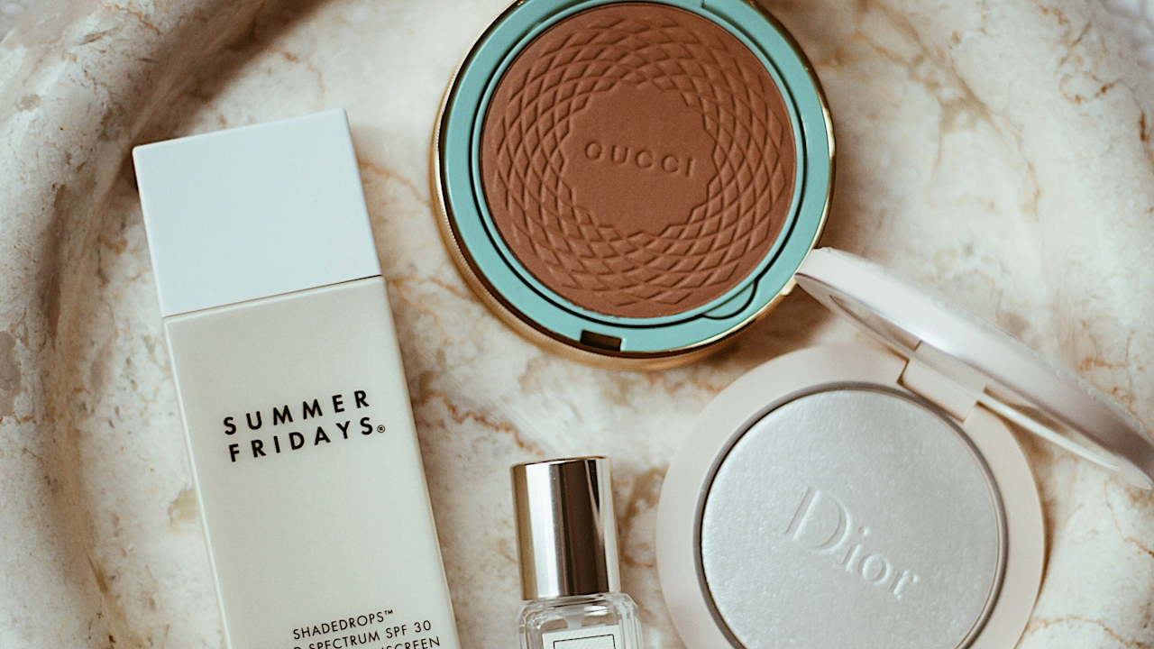 A collection of various makeup products displayed indoors, including OUGDI Summer Fridays and Dior ShadeCrops SPFs. The scene emphasizes a tan powder among other cosmetics, reflecting a fashion aesthetic.