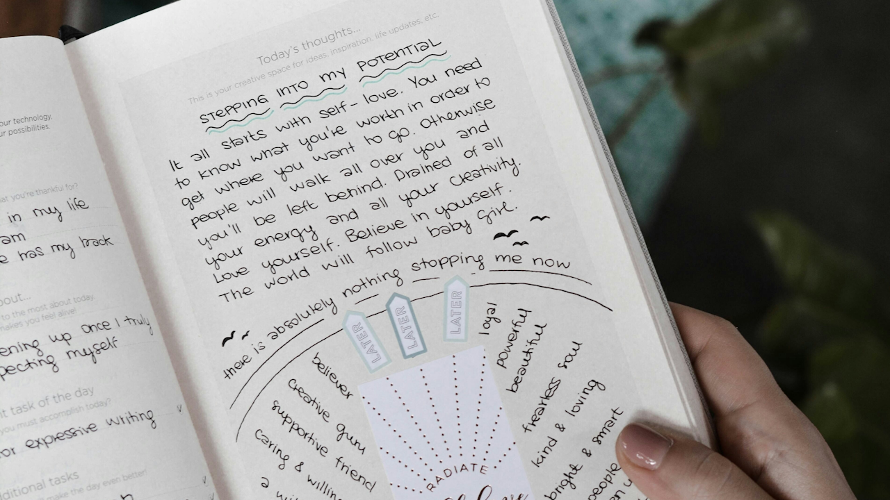 A hand holding a book titled "Stepping Into My Potential," which contains motivational text about self-love, worth, and personal growth. The book's pages feature colorful ink and handwritten notes emphasizing the importance of believing in oneself and embracing creativity.