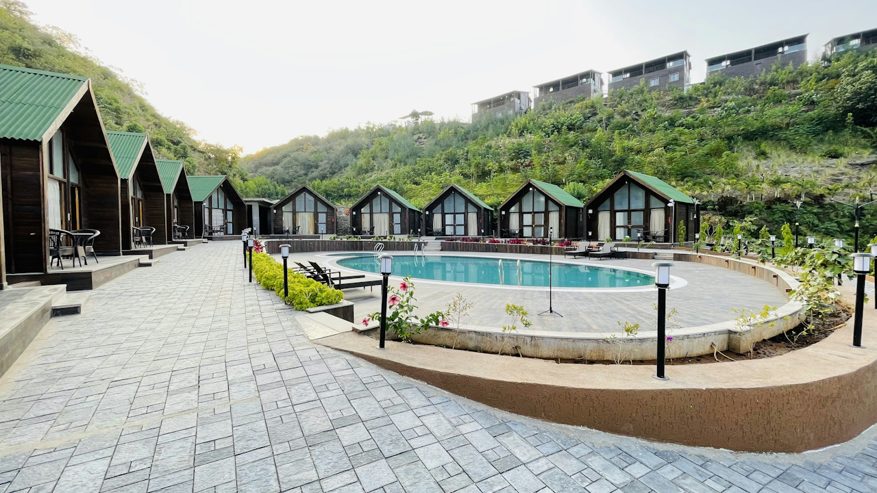 A panoramic view of an outdoor swimming pool surrounded by lush greenery and trees, with a backdrop of a resort building under a clear sky. The pool is part of a spa town, featuring water and plant elements in the landscape.