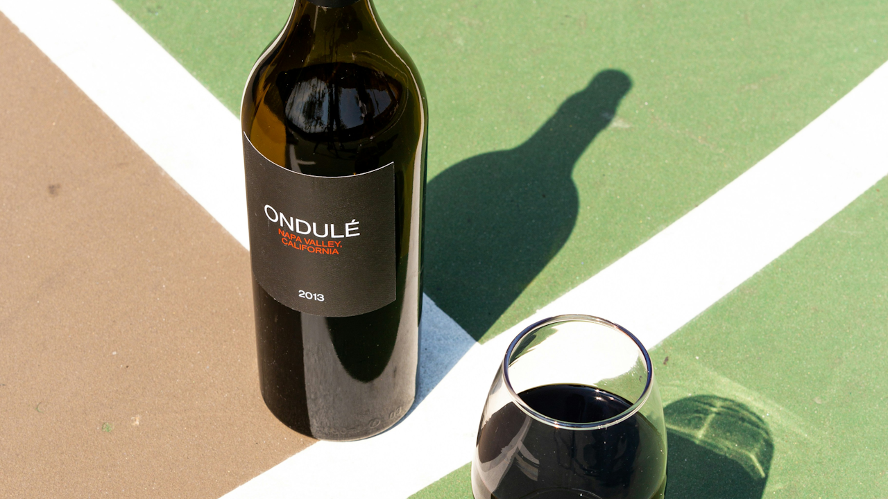 Bottle of 2013 Ondulé Napa Valley wine and glass on green surface with white lines, casting shadows in sunlight.