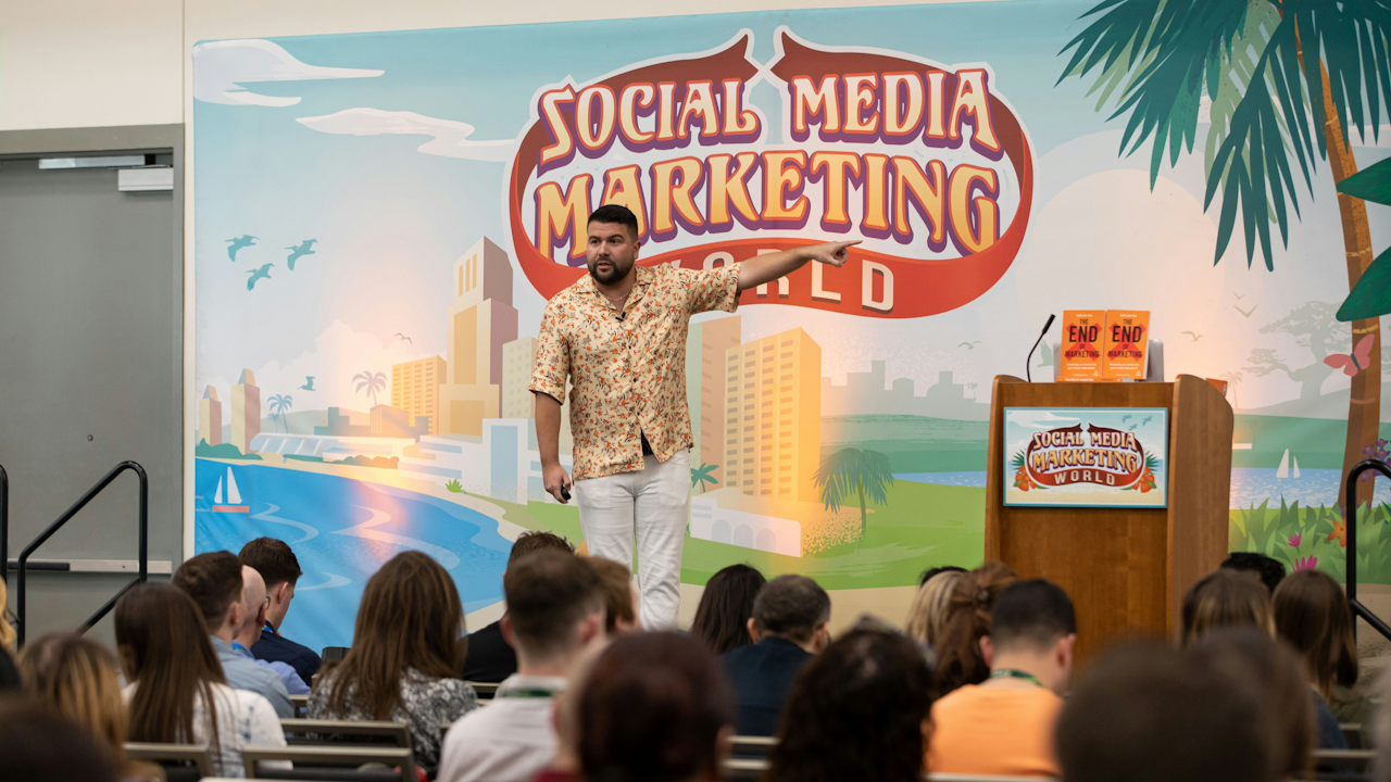 Speaker in floral shirt presenting at Social Media Marketing World event, with audience and colorful beach-themed backdrop.