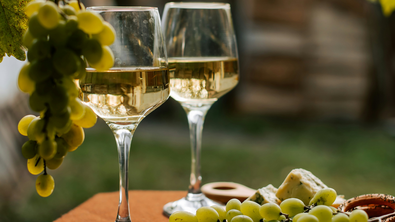 Two wine glasses filled with white wine are on a table next to a bunch of green grapes and assorted cheeses, with a blurred outdoor background.